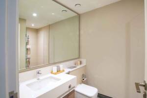 Clean modern bathroom with a large mirror, white sink, beige tiles, and shower area visible through the reflection. Minimalist design with neutral tones and organized accessories.
