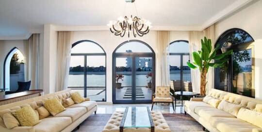 Luxurious living room with panoramic waterfront views, elegant furniture, and stylish decor featuring a large potted plant and modern chandelier.