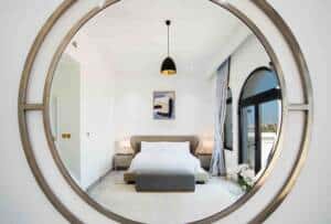 Reflective metallic round mirror showing modern bedroom with a bed, bedside tables, abstract art, and large arched window with view of cityscape, interior designer decor, and minimalist style.