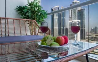Luxurious balcony with glass railing overlooking Dubai skyline, featuring a table with wine glasses, fresh fruit, and a potted plant, perfect for relaxing and enjoying stunning city views.