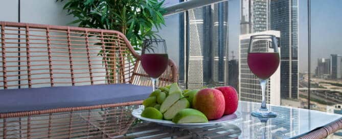 Luxurious balcony with glass railing overlooking Dubai skyline, featuring a table with wine glasses, fresh fruit, and a potted plant, perfect for relaxing and enjoying stunning city views.